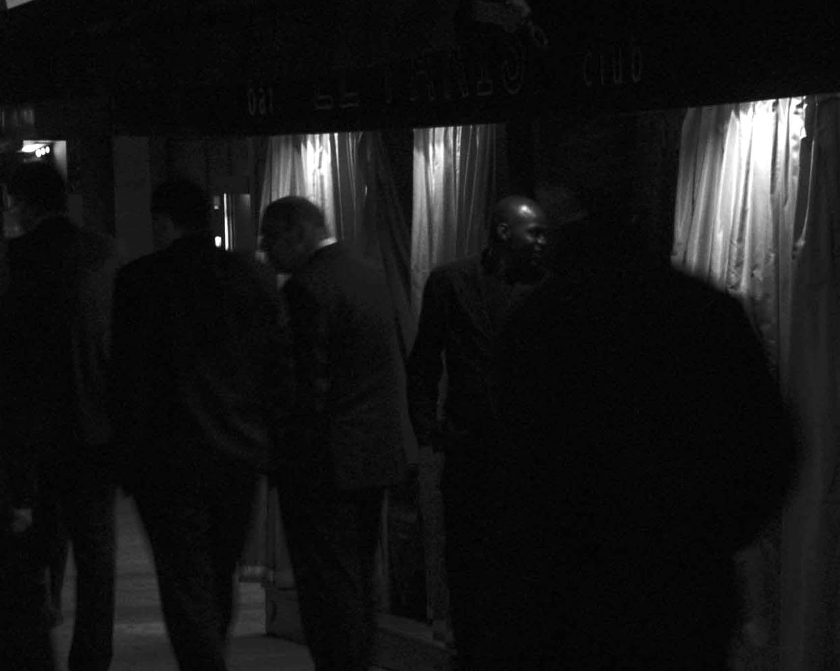 Men in dark suits outside a club entrance with lit curtains