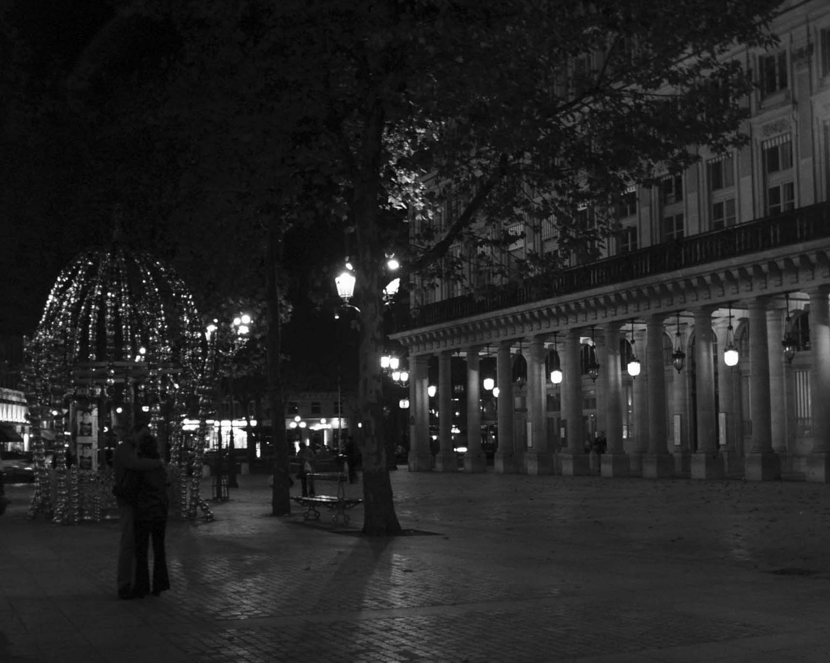 Couple embracing beside a lit kiosk, Place Colette and the Comédie-Française arcades