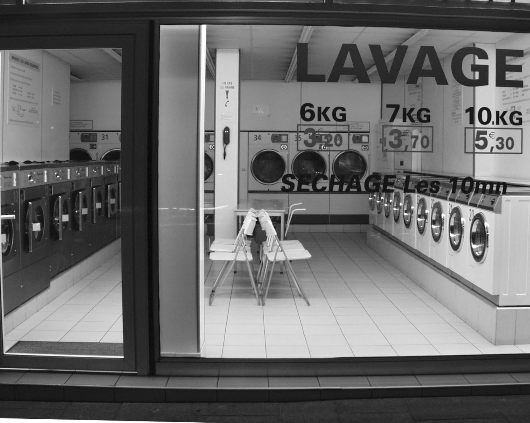 Empty launderette seen through its window, lettered Lavage with prices