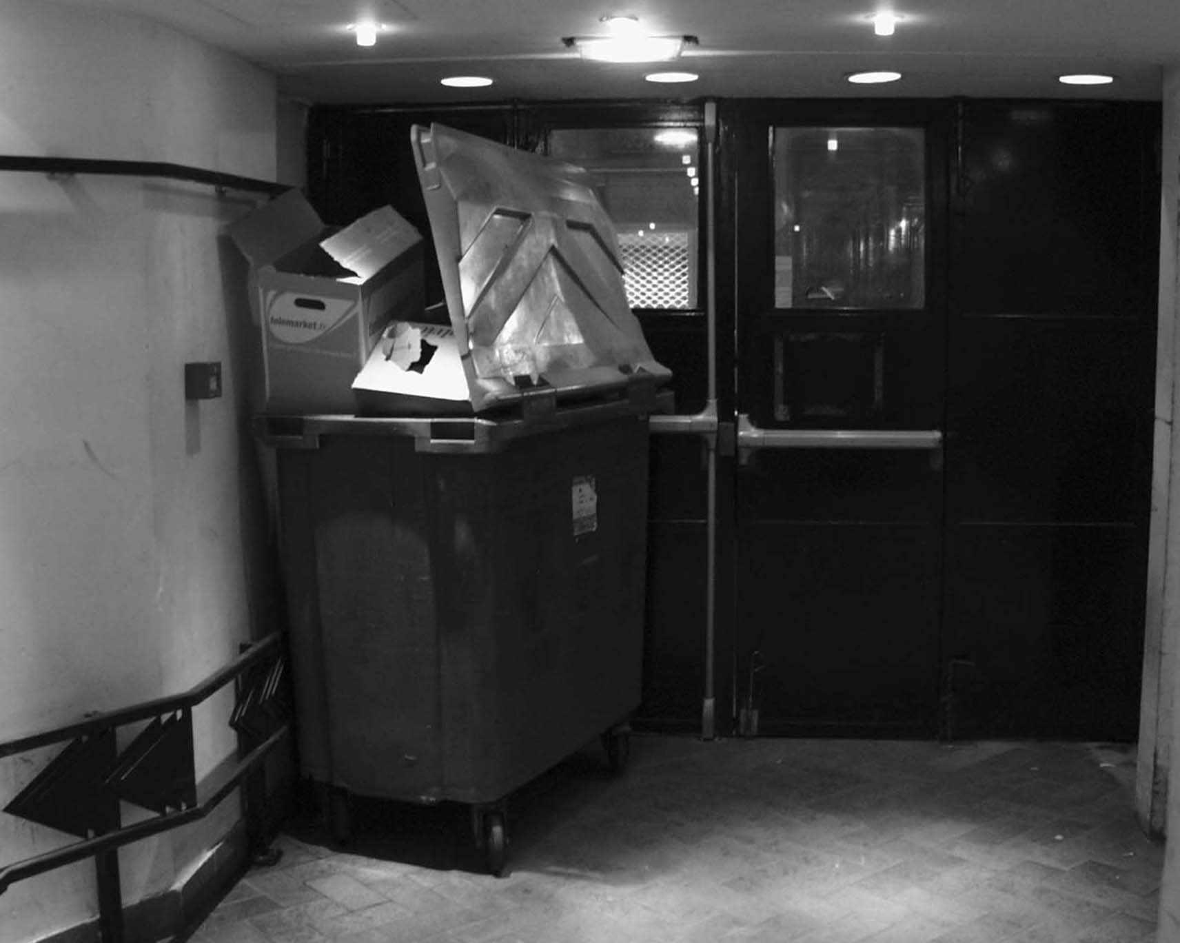 Overflowing bin beside black service doors in a basement corridor