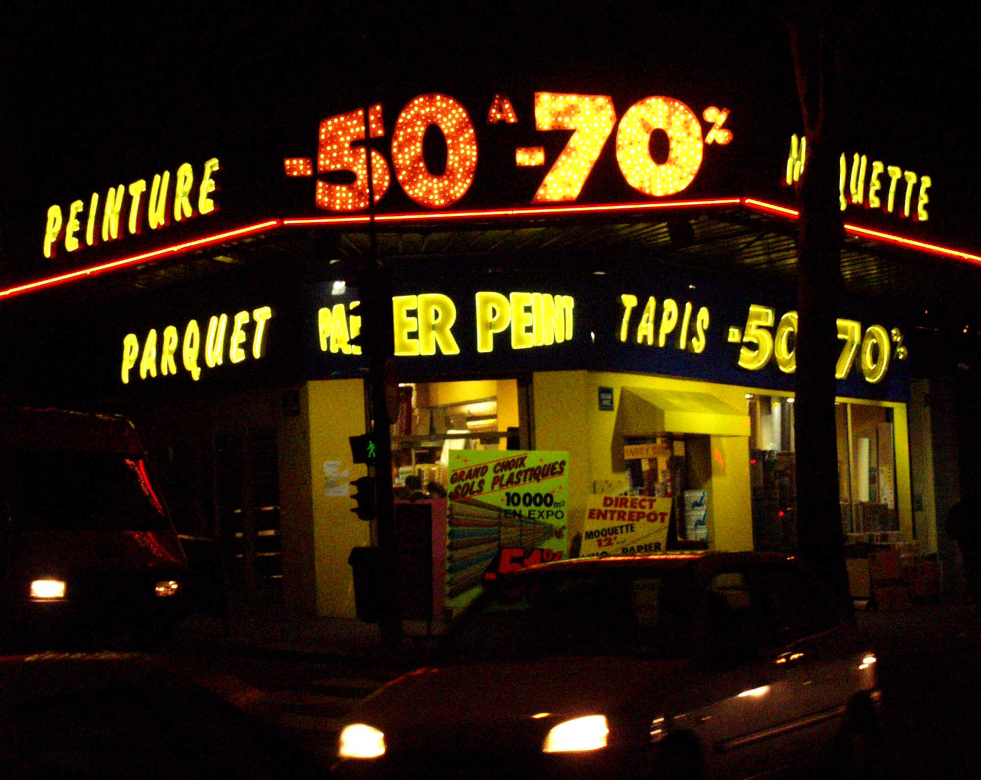 Shop at night with yellow neon reading Peinture, Parquet, Tapis, -50 -70%