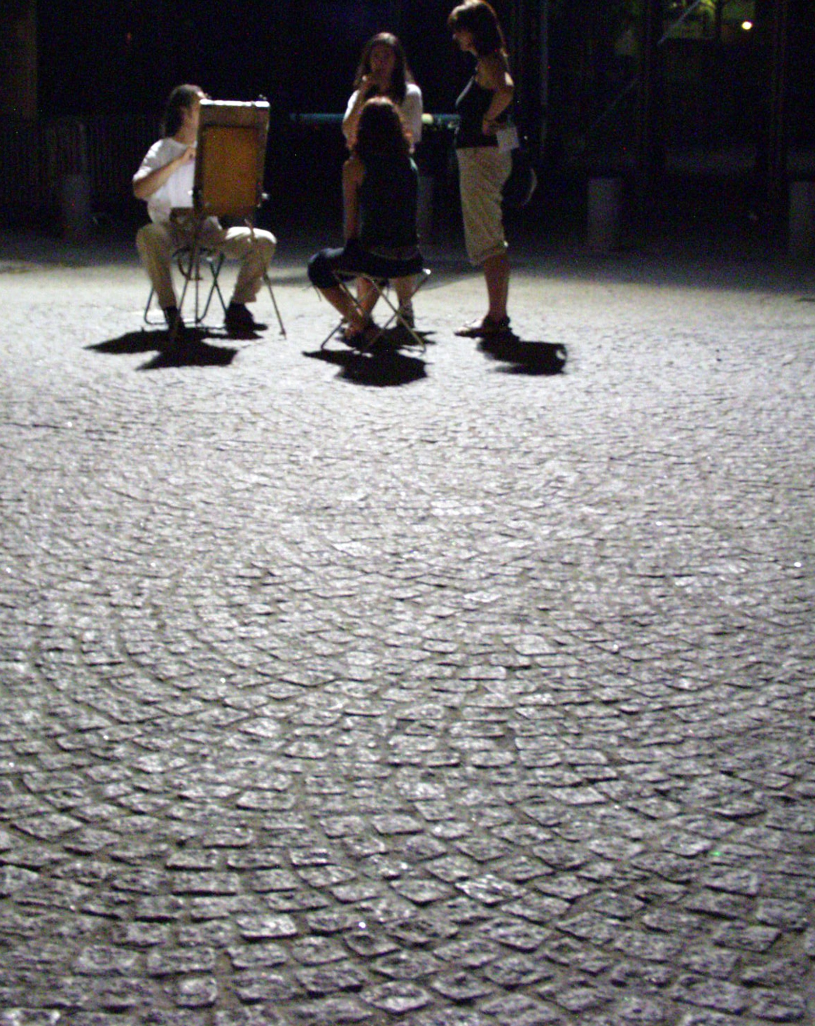 Cobbles under a streetlight, three people sitting on folding chairs in the dark