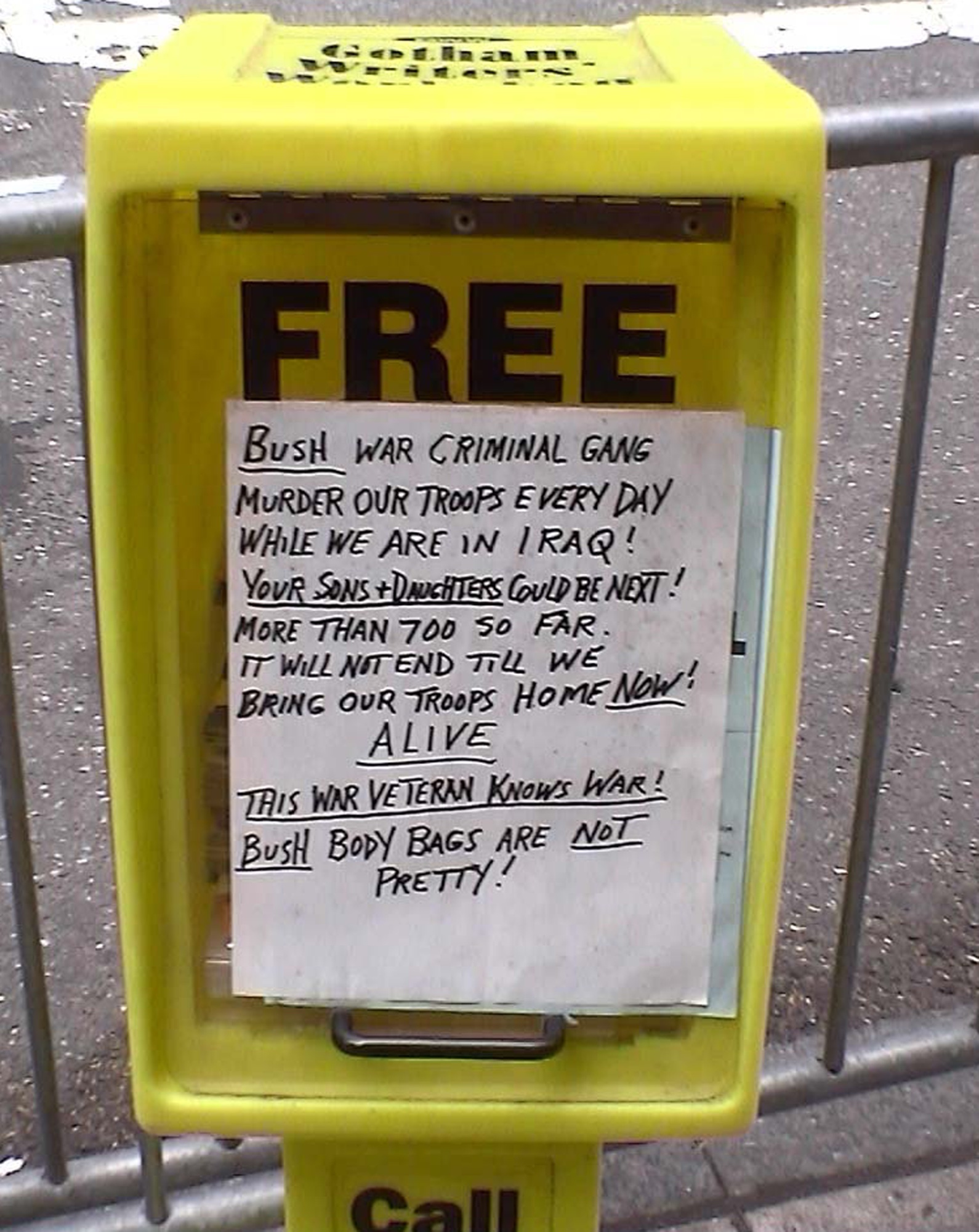 Yellow newspaper box reading Free, a handwritten anti-war note taped to it