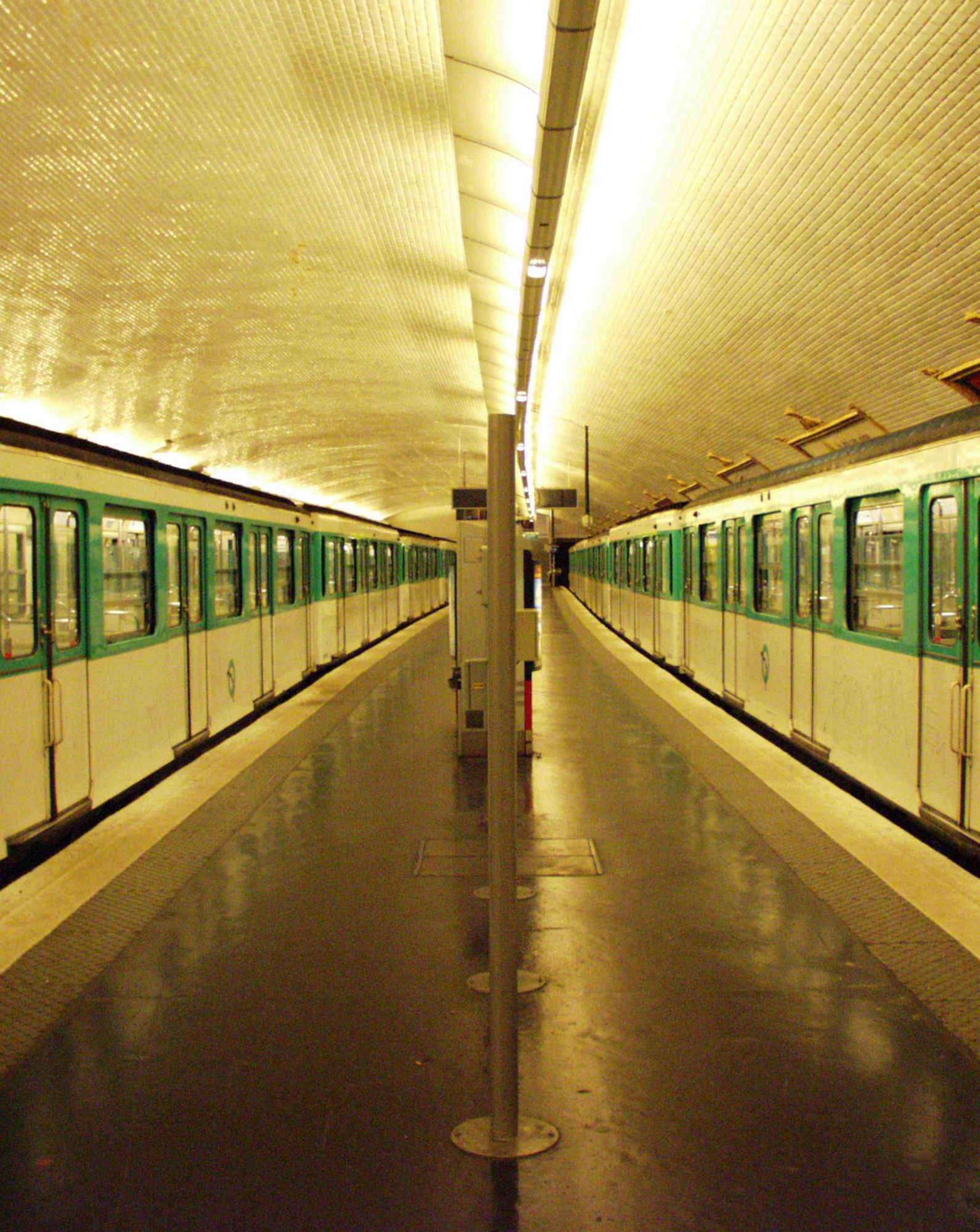 Empty métro platform between two green and white trains, yellow vaulted tiles