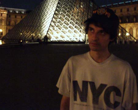 The young man in his NYC T-shirt and cap beside the lit Louvre pyramid